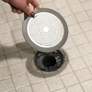 Graues rundes Haarsieb mit gelochtem Metalleinsatz wird von einer Hand über einem Bodenablauf gehalten