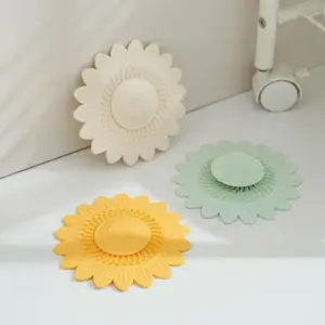 Drei Abflussabdeckungen in Sonnenblumenform in Cremeweiß, Mintgrün und Gelb liegen auf einem weißen Untergrund