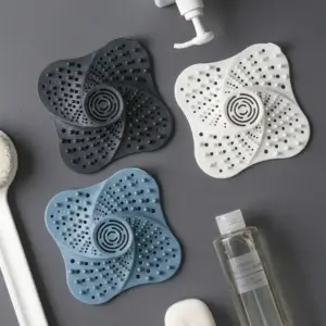 Drei geschwungene Abflusssiebe in Schwarz, Weiß und Blau mit Lochmuster auf grauem Untergrund neben Badutensilien
