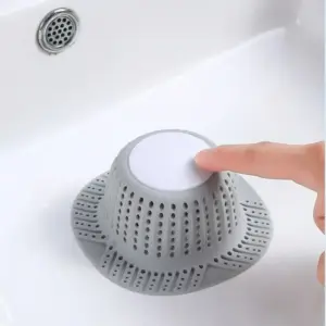 Graues hutförmiges Abflusssieb mit weißer Mitte in einem Waschbecken, ein Finger drückt darauf