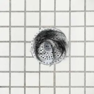 Weißes Abflusssieb mit Lochmuster liegt im gefliesten Boden und fängt dunkle Haare auf